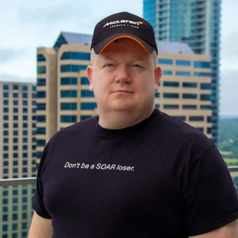 A man wearing a black McLaren hat and a black t-shirt that says Dont be a SOAR loser. stands on a balcony with tall office buildings in the background.