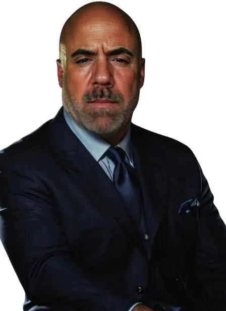 A bald man with a trimmed beard wears a dark blue suit, matching tie, and pocket square, posing against a plain white background with a serious expression.