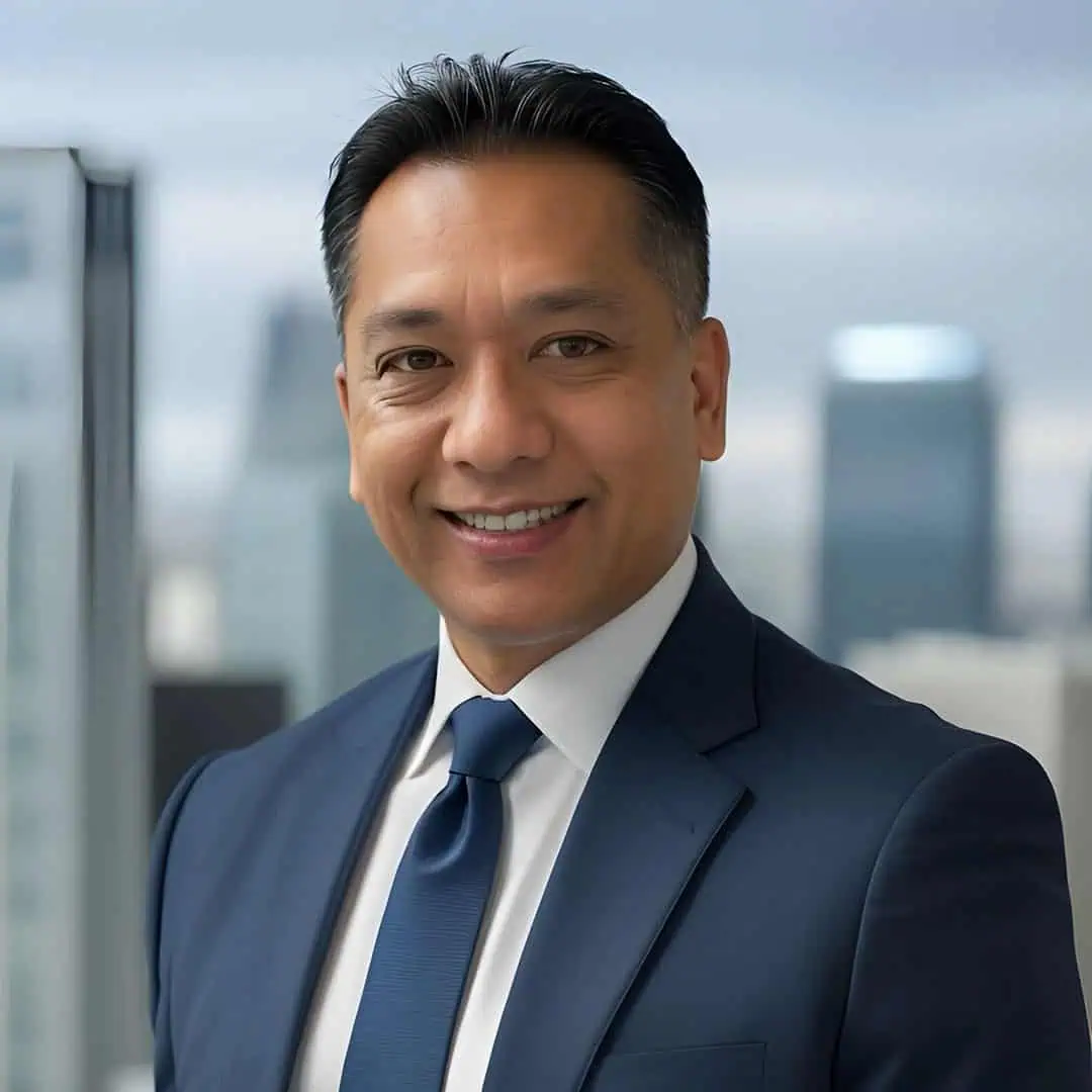 A man in a navy suit and tie smiles while standing in an office with large windows and a cityscape in the background.