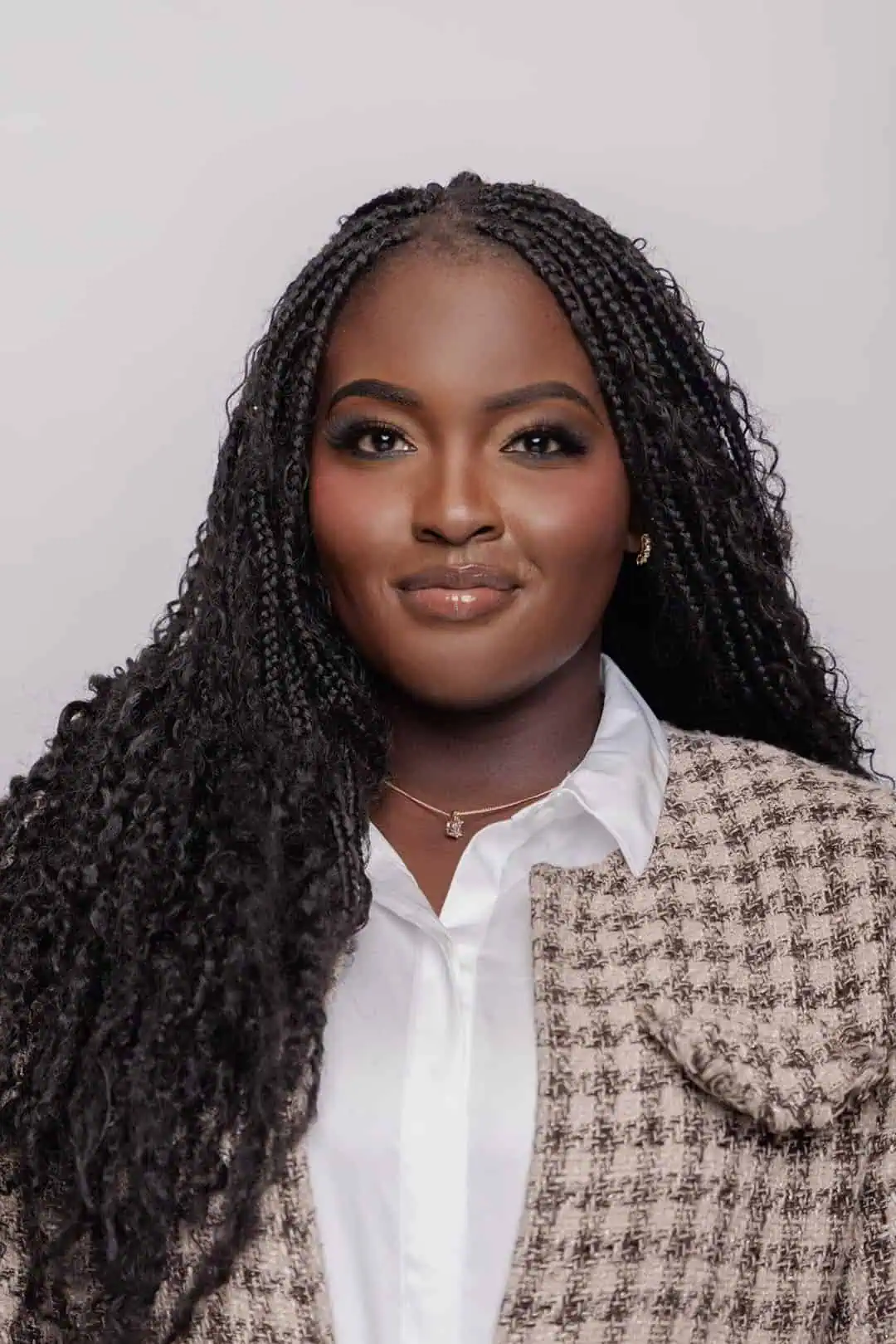 A woman with long, braided hair smiles softly. She wears a white collared shirt under a plaid jacket and a delicate necklace. The background is plain and light-colored.