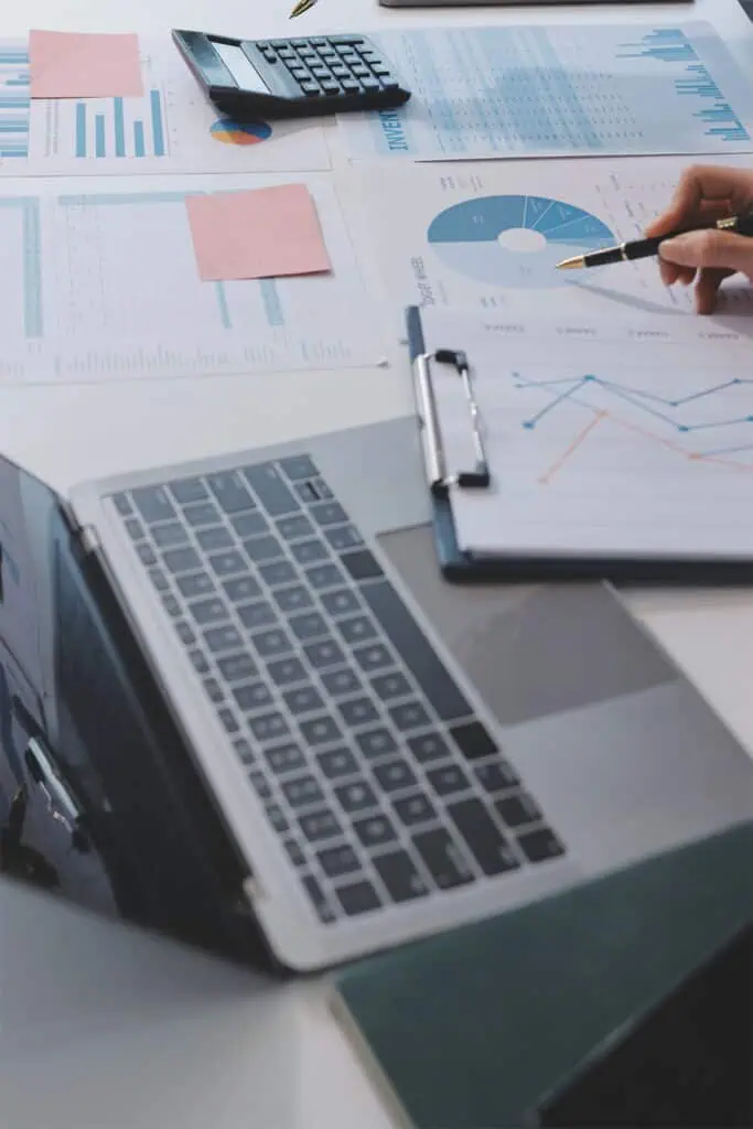 A person points at a clipboard with charts, surrounded by a laptop, calculator, documents with graphs, and pink sticky notes on a desk, suggesting a work or business analysis setting.