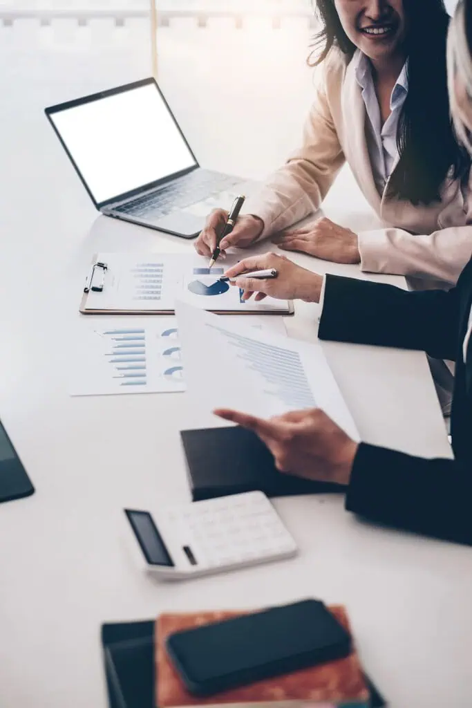 Two people in business attire discussing financial charts and graphs at a desk with a laptop, papers, calculator, and clipboard, focusing on a pie chart document.