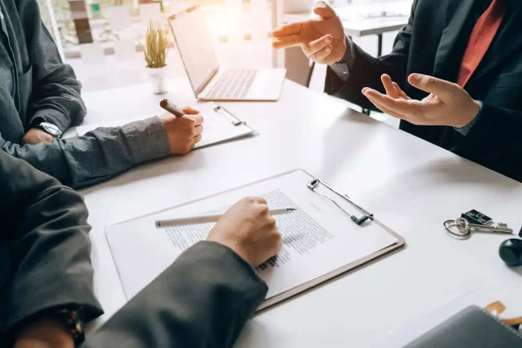 Three people sit at a table in a business meeting, with documents, a clipboard, a laptop, and keys on the desk. One person gestures while others take notes. A small plant is in the background.
