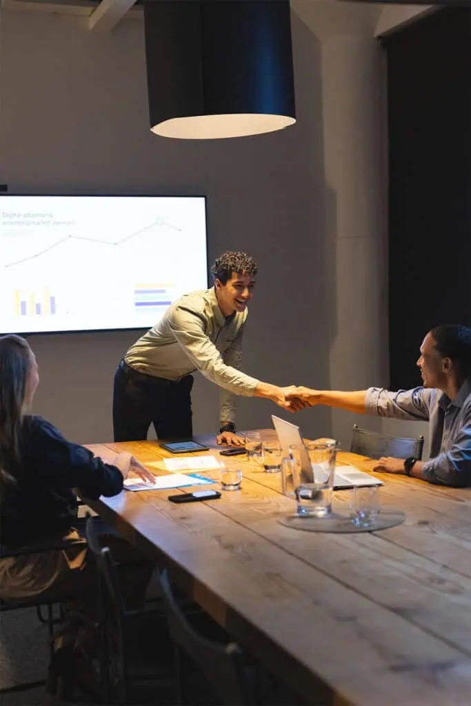 Three people sit at a conference table in a meeting room. One person stands, leaning forward to shake hands with another seated person. A presentation with charts is displayed on a screen in the background.