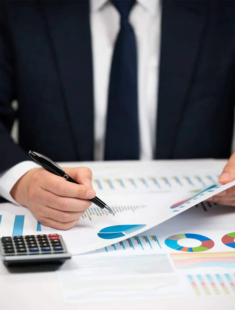 A person in a suit reviews financial charts and graphs on paper, holding a pen and a document. A calculator is placed on the desk, along with various colorful data visualizations.