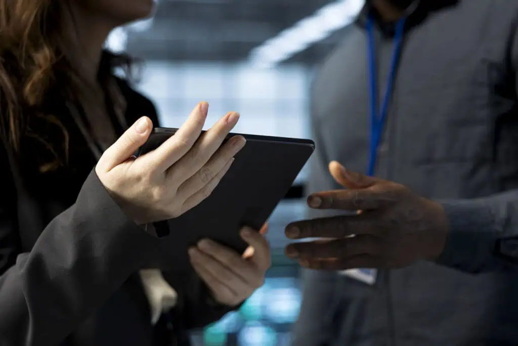 Two people stand indoors, one holding a tablet and gesturing, while the other listens with hands partially visible. Both are dressed in business attire, suggesting a professional or collaborative setting.