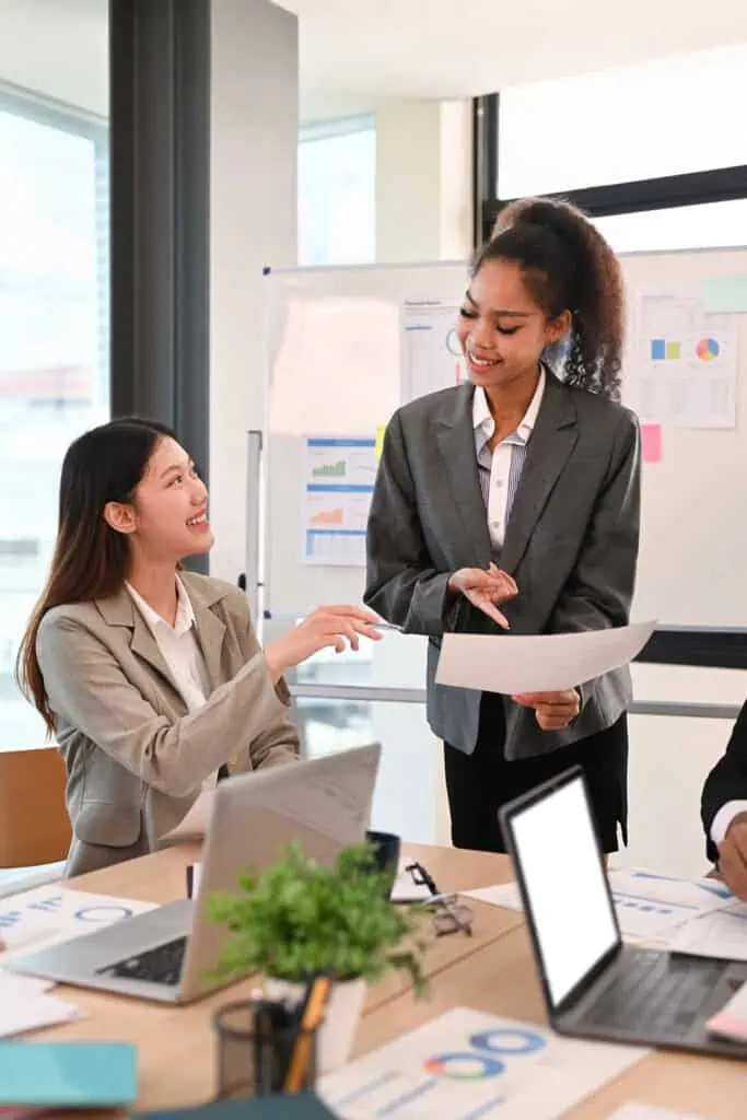 Two businesswomen in suits smiling and discussing documents in a modern office, with laptops, paperwork, and charts visible on a table and a whiteboard with graphs in the background.