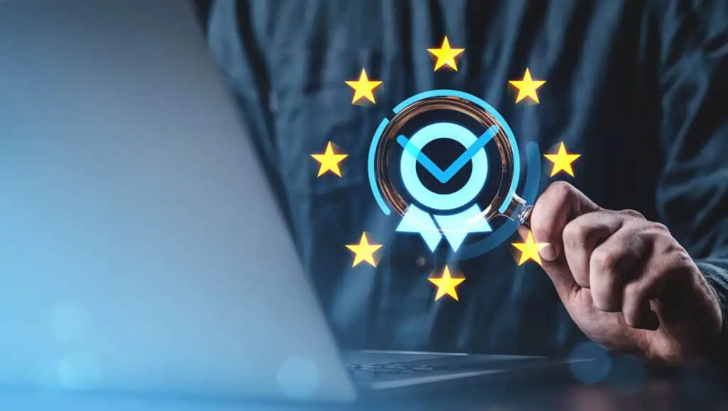 A person uses a magnifying glass near a laptop, with a digital checkmark badge surrounded by yellow stars, symbolizing data protection and compliance, likely referencing the European Unions GDPR.