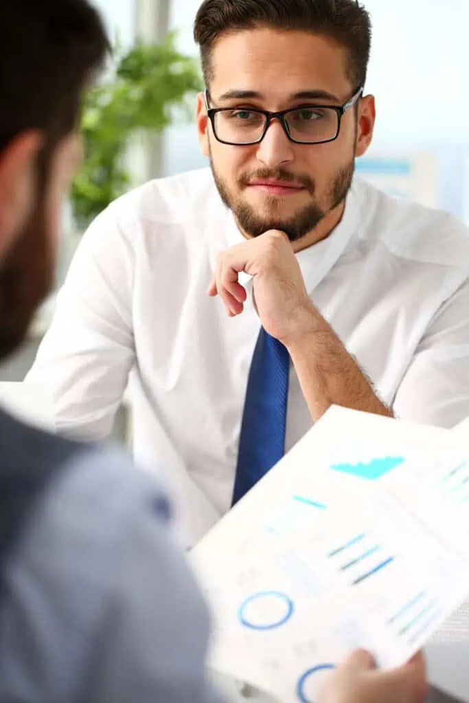 A man with glasses and a beard, wearing a white shirt and blue tie, listens attentively to another person holding a paper with blue charts and graphs during a business meeting.