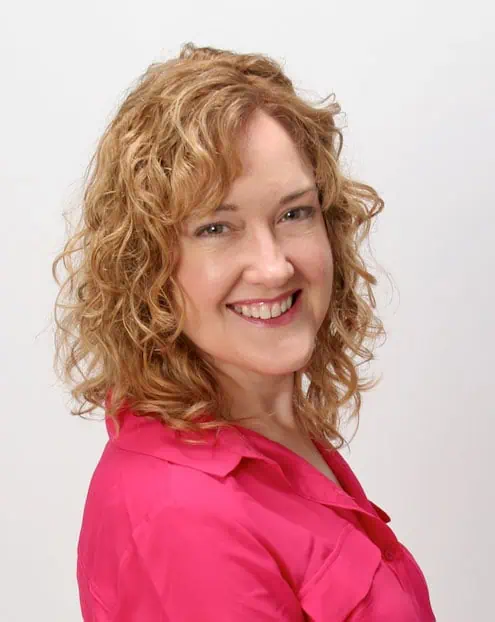 A woman with curly, shoulder-length blonde hair smiles at the camera, wearing a bright pink blouse against a plain white background.