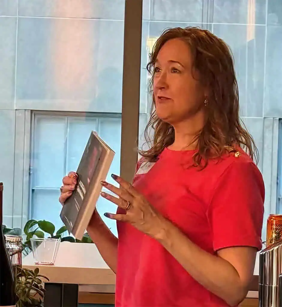 A woman with wavy brown hair wearing a red shirt stands indoors, holding up a book or magazine while speaking. She appears to be addressing a group, with a window and plants visible in the background.