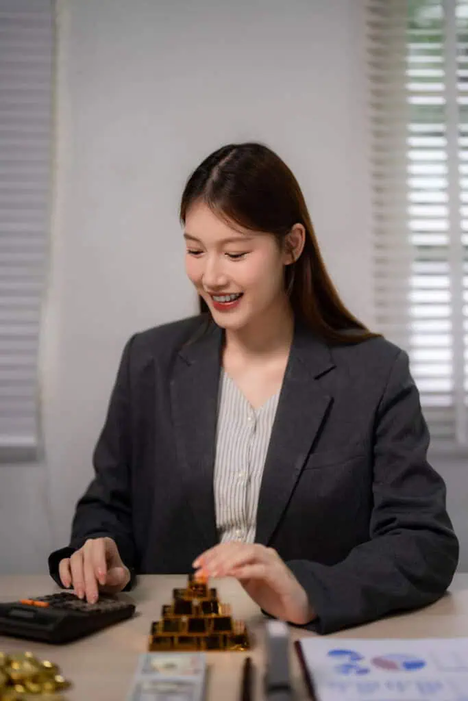 A woman in a dark blazer sits at a desk, smiling while using a calculator and stacking small gold bars. Documents and cash are visible on the desk in front of her.
