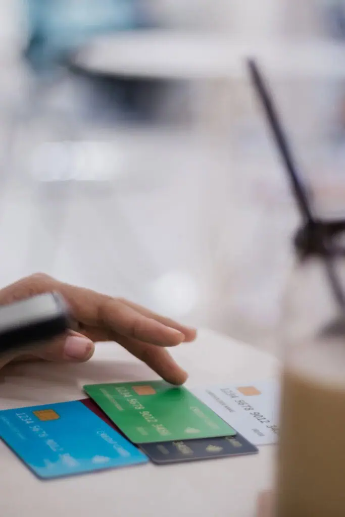 A hand hovers over three credit cards—blue, green, and white—on a table, with a blurred drink and straw in the foreground.