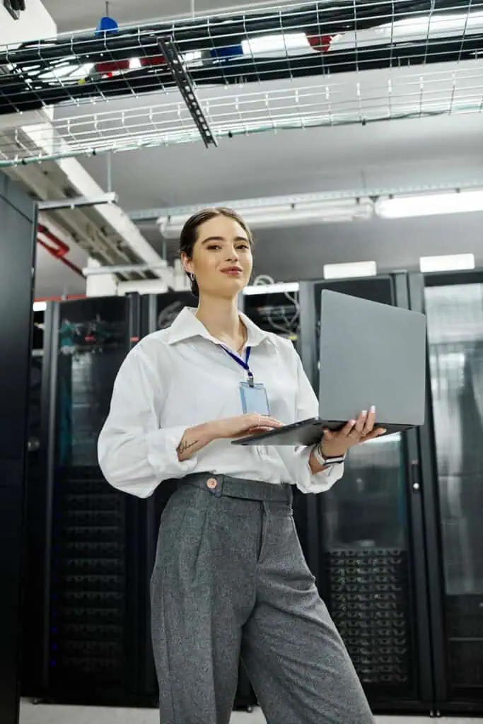 A woman in business attire stands in a server room holding an open laptop, looking confidently at the camera. She wears a white shirt, gray pants, and a lanyard with an ID badge. Server racks are visible in the background.