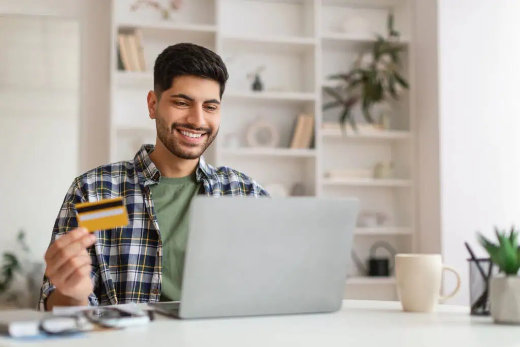 A smiling man sits at a desk holding a credit card in one hand and using a laptop with the other. Shelves, a mug, and plants are visible in the background, suggesting a home office setting.
