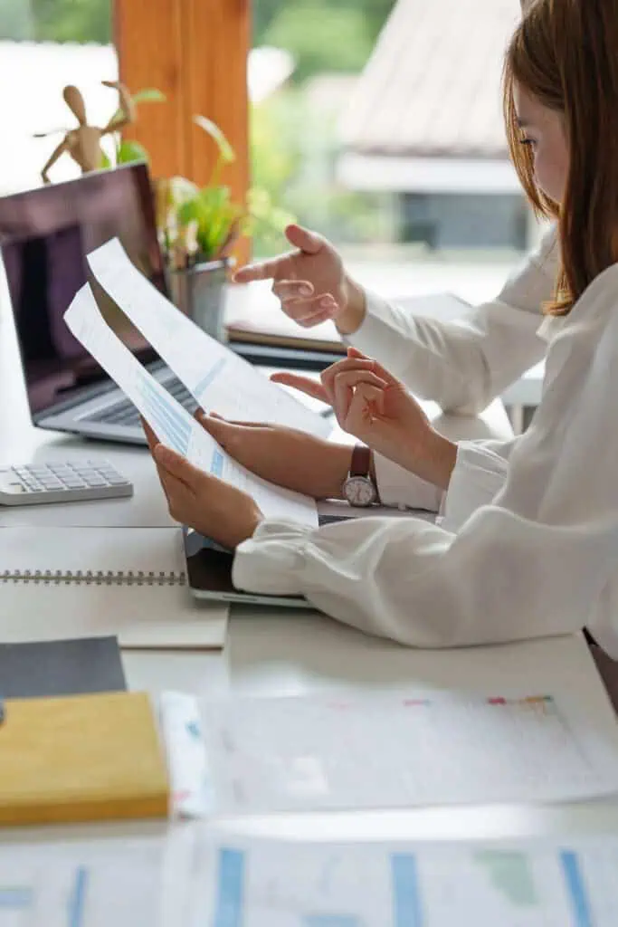 Two people sitting at a desk reviewing printed charts or documents together, with a laptop, notebook, and papers spread out in front of them in a bright office setting.
