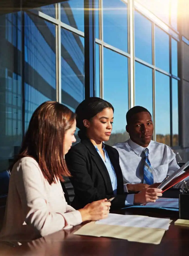 Three business professionals sit at a table in a modern office with large windows, discussing documents together. Sunlight streams in, illuminating their focused expressions.