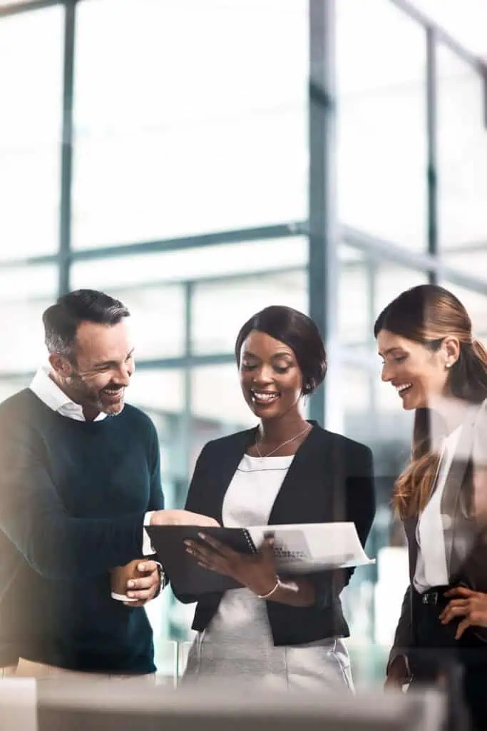 Three business professionals stand together in a modern office, smiling and looking at documents on a clipboard. One person points at the clipboard while the others listen and engage in discussion.