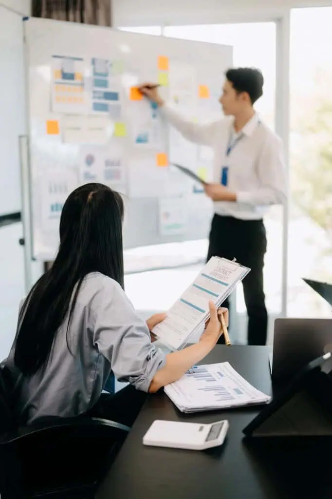 A woman holding charts sits at a desk, analyzing documents, while a man in business attire stands and writes on a whiteboard covered with graphs and sticky notes in a bright office.