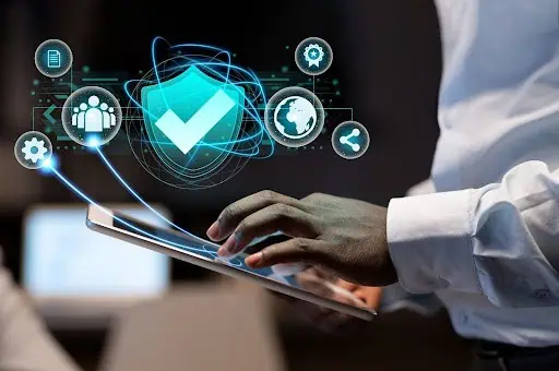 A person using a tablet device with digital security icons and a large checkmark shield graphic floating above it, representing cybersecurity, data protection concepts, and SOC 2 compliance.